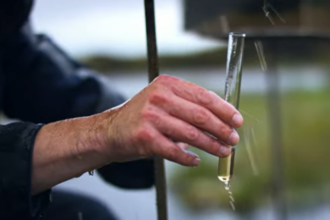 Experiment showing a hand holding a small glass tube collecting a sample of water dripping from a bucket of peat.