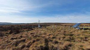 Peatland landscape showing flux tower and solar panel with a blue sky and hills in the background. Image credit Hollie Cooper, UKCEH