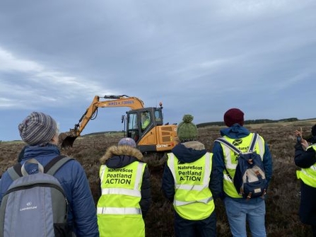 Group of people from NatureScot Peatland ACTION watch a machine operator installing peat dams along a peatland ditch.